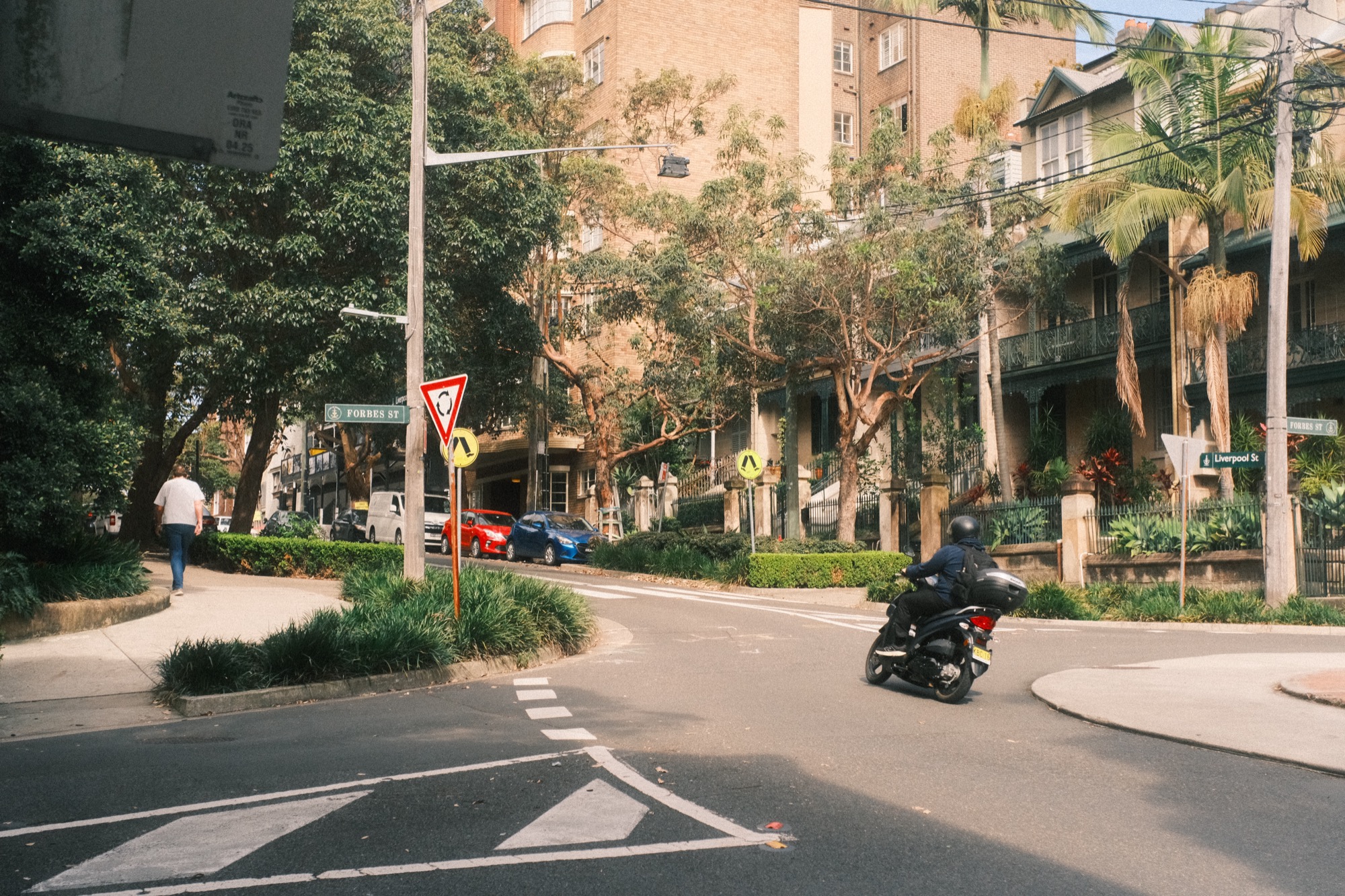 At a junction, a man in a white shirt walks away from the camera; a scooter crosses behind him.