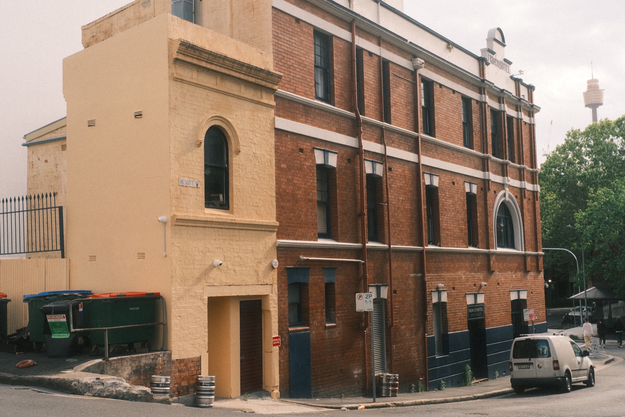 Kegs stand by a back door behind a brick building; a white van is parked beside them.