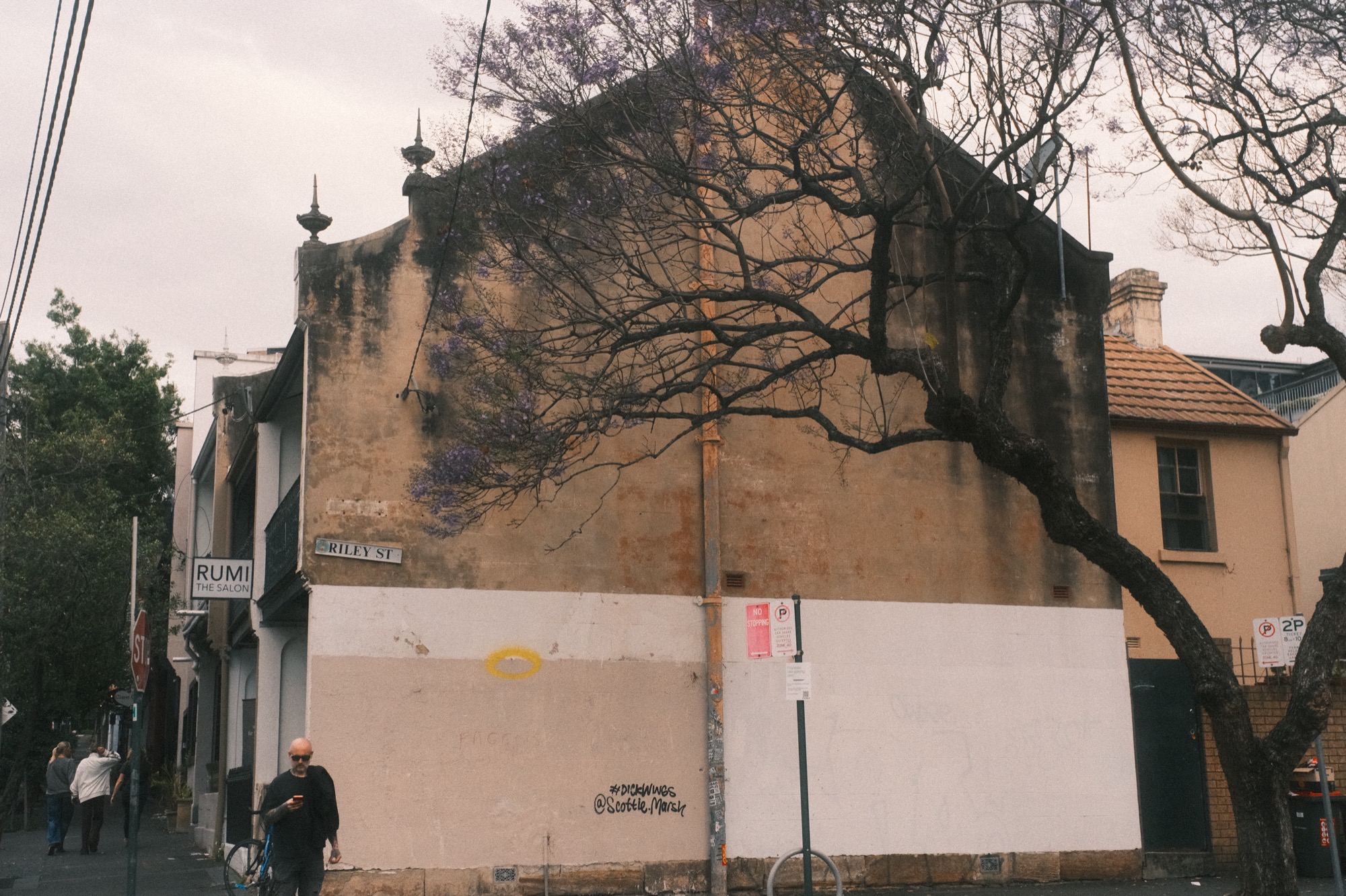 The lower half of a gable wall is painted over in a flat pale colour; a yellow ring and a scrawl sit on the paint. A man walks past.